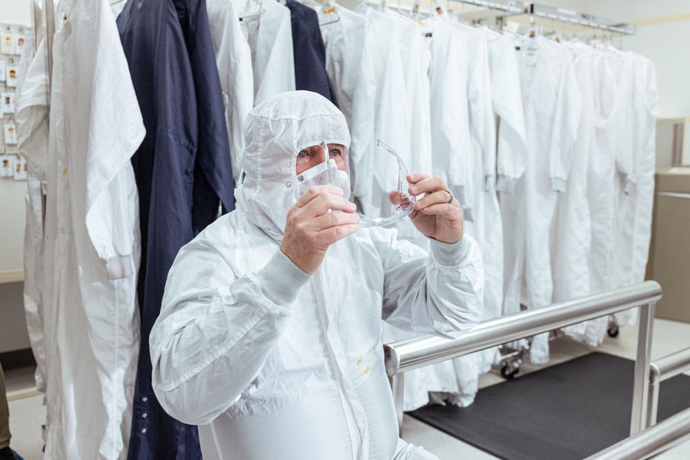 A person wearing a full-body white protective suit and hood puts on safety goggles in a cleanroom, highlighting semiconductor manufacturing careers and workforce development in the high-tech industry.