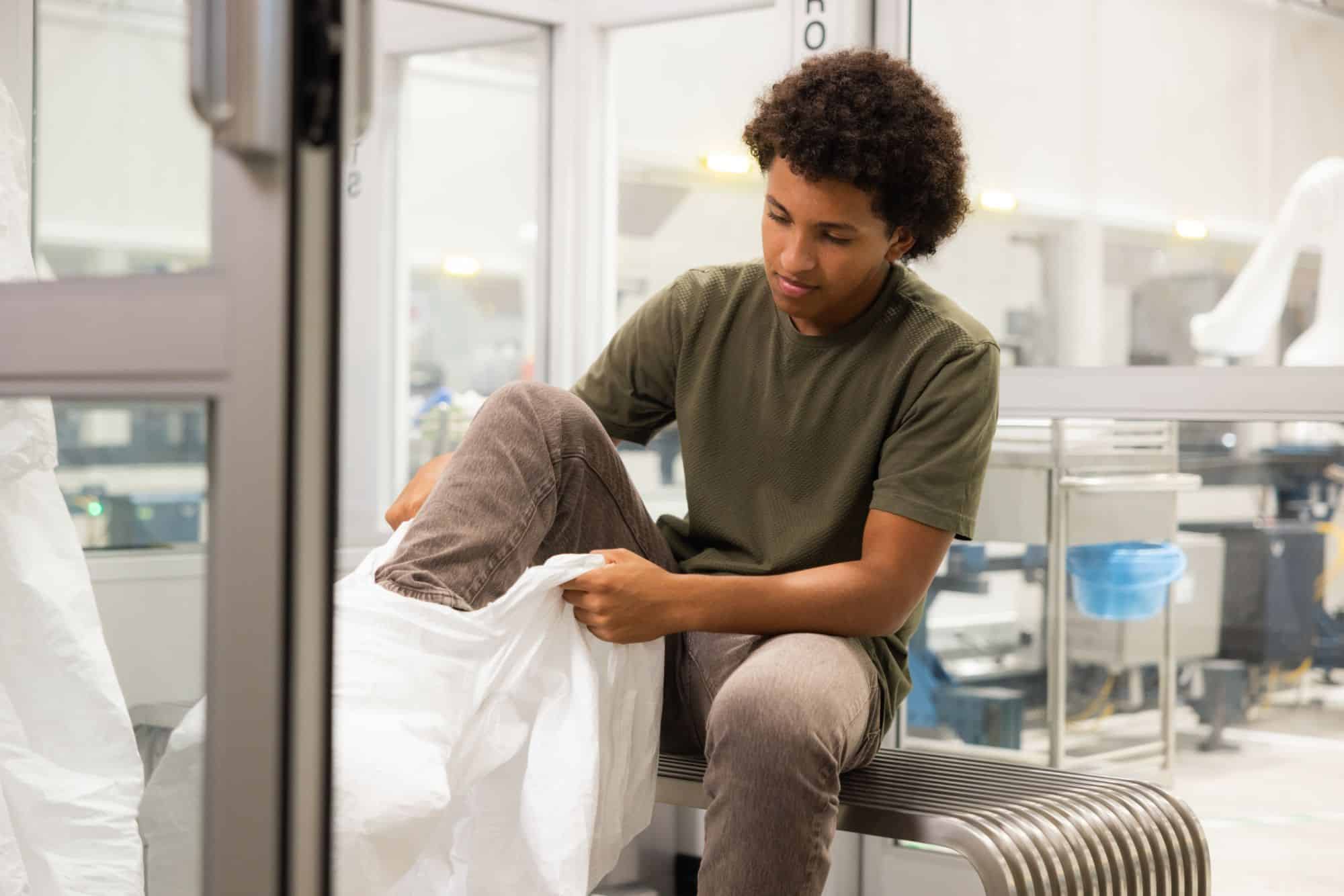 A young person sits on a bench, wearing a green shirt and brown pants, while putting on a white protective suit in a clean, modern laboratory—preparing to explore semiconductor engineering careers and exciting tech opportunities for students.